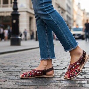 May include: Red, blue, and yellow embroidered sandals with brown leather straps and a light-colored sole. The sandals are worn with blue jeans with frayed hems, walking on a cobblestone street.