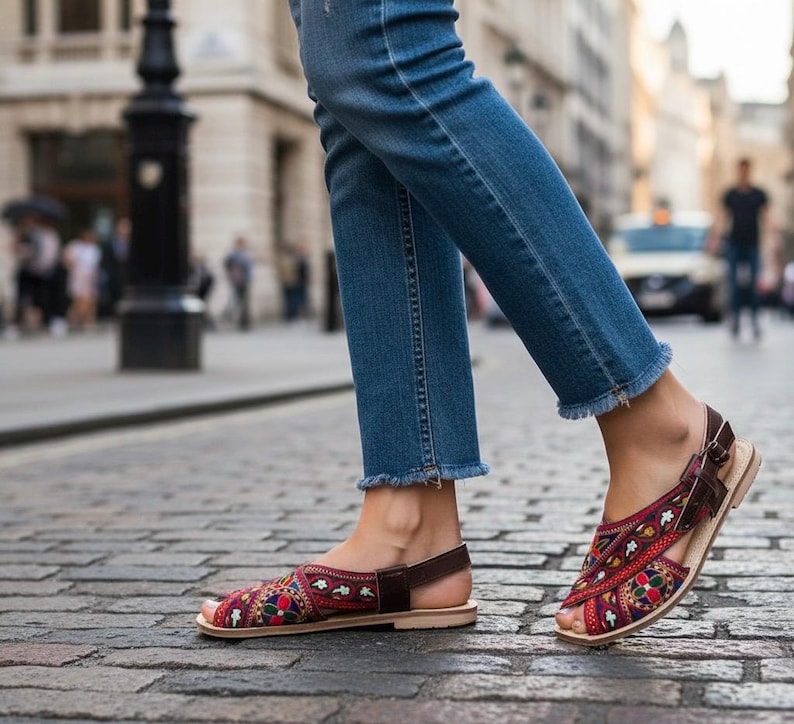 May include: Red, blue, and yellow embroidered sandals with brown leather straps and a light-colored sole. The sandals are worn with blue jeans with frayed hems, walking on a cobblestone street.