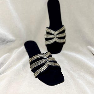 May include: A pair of black velvet slip-on sandals. The sandals feature a square toe and a decorative strap across the top, embellished with silver-colored beads. The background is a light, neutral fabric.