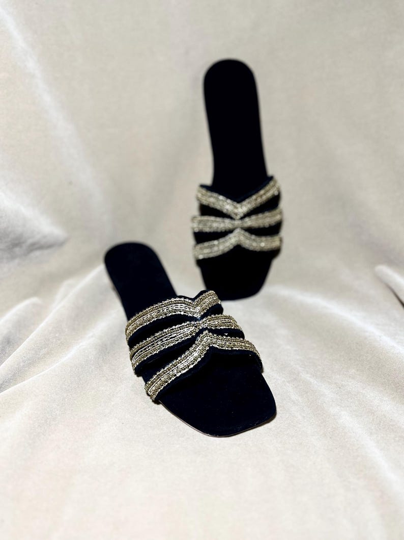 May include: A pair of black velvet slip-on sandals. The sandals feature a square toe and a decorative strap across the top, embellished with silver-colored beads. The background is a light, neutral fabric.