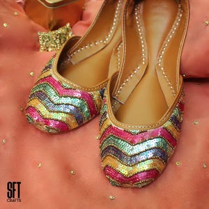 May include: A pair of colorful, sequined flats with a chevron pattern. The shoes feature a tan leather insole and a decorative design in shades of pink, yellow, green, and purple. The shoes are displayed on a peach-colored surface.
