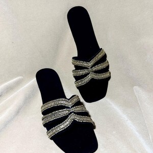 May include: A pair of black velvet slide sandals. The sandals feature a bow-like design across the top, embellished with rows of silver beads. The open-toe design and flat sole provide a comfortable and stylish look.