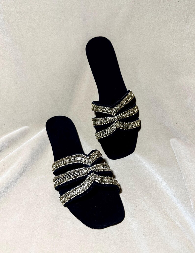 May include: A pair of black velvet slide sandals. The sandals feature a bow-like design across the top, embellished with rows of silver beads. The open-toe design and flat sole provide a comfortable and stylish look.