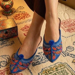 May include: A pair of blue embroidered slip-on shoes with a pointed toe and a small blue pom-pom detail. The shoes feature colorful floral and geometric patterns in red, orange, and green. The shoes are on a patterned rug.