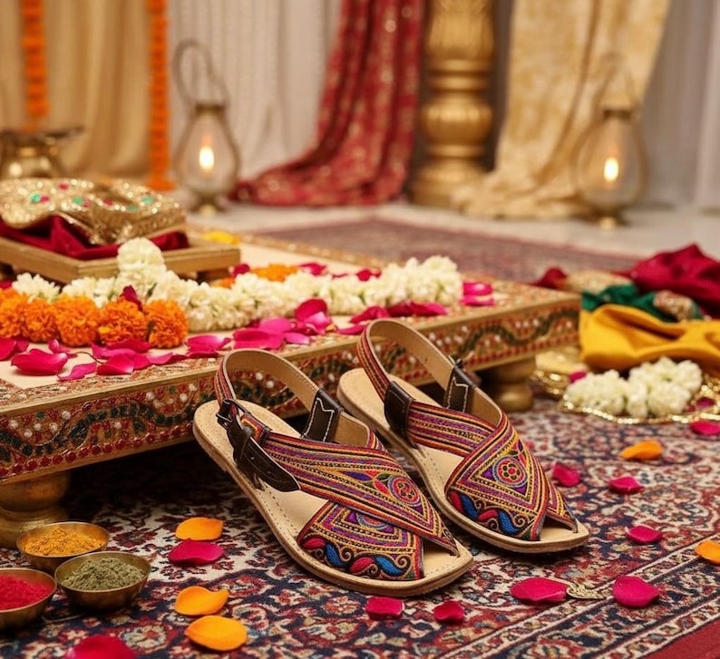 May include: A pair of colorful, patterned sandals with brown leather straps. The sandals feature intricate embroidery in shades of red, purple, blue, and yellow. They are displayed on a decorative table with floral arrangements and rose petals.