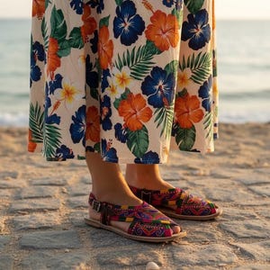 May include: Colorful, patterned sandals with a leather strap and buckle. The sandals are worn on a sandy beach, paired with a floral print skirt featuring orange, blue, and white flowers.