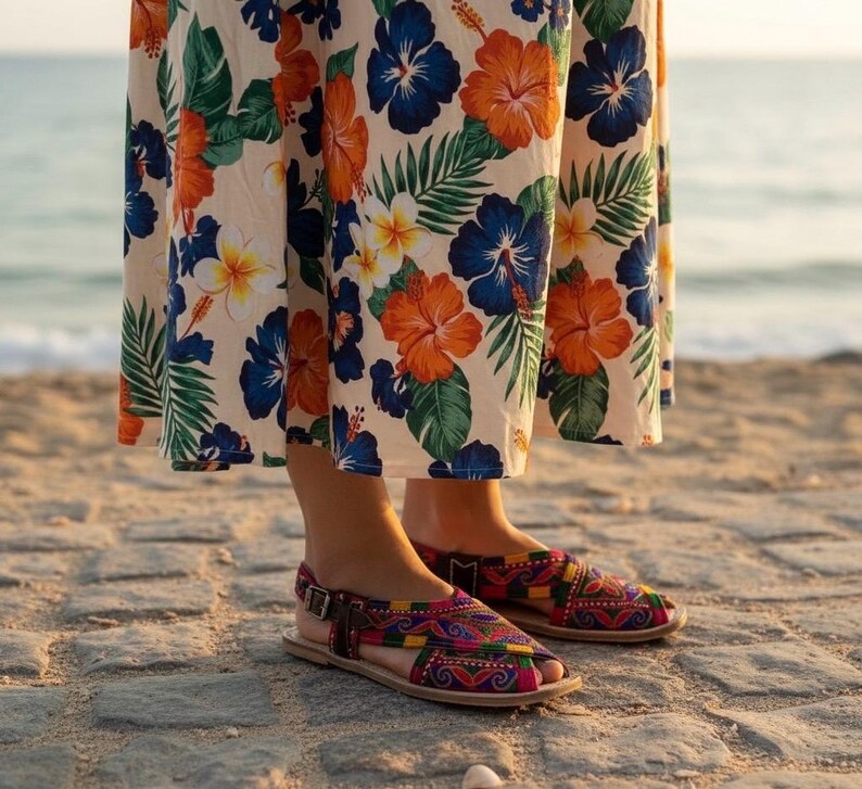 May include: Colorful, patterned sandals with a leather strap and buckle. The sandals are worn on a sandy beach, paired with a floral print skirt featuring orange, blue, and white flowers.