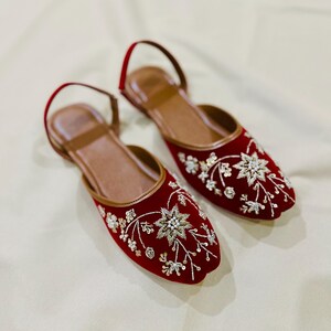 May include: A pair of red velvet flats with intricate floral embroidery in silver and gold. The shoes have a brown leather insole and a thin ankle strap. The flats are a stylish and elegant footwear option.