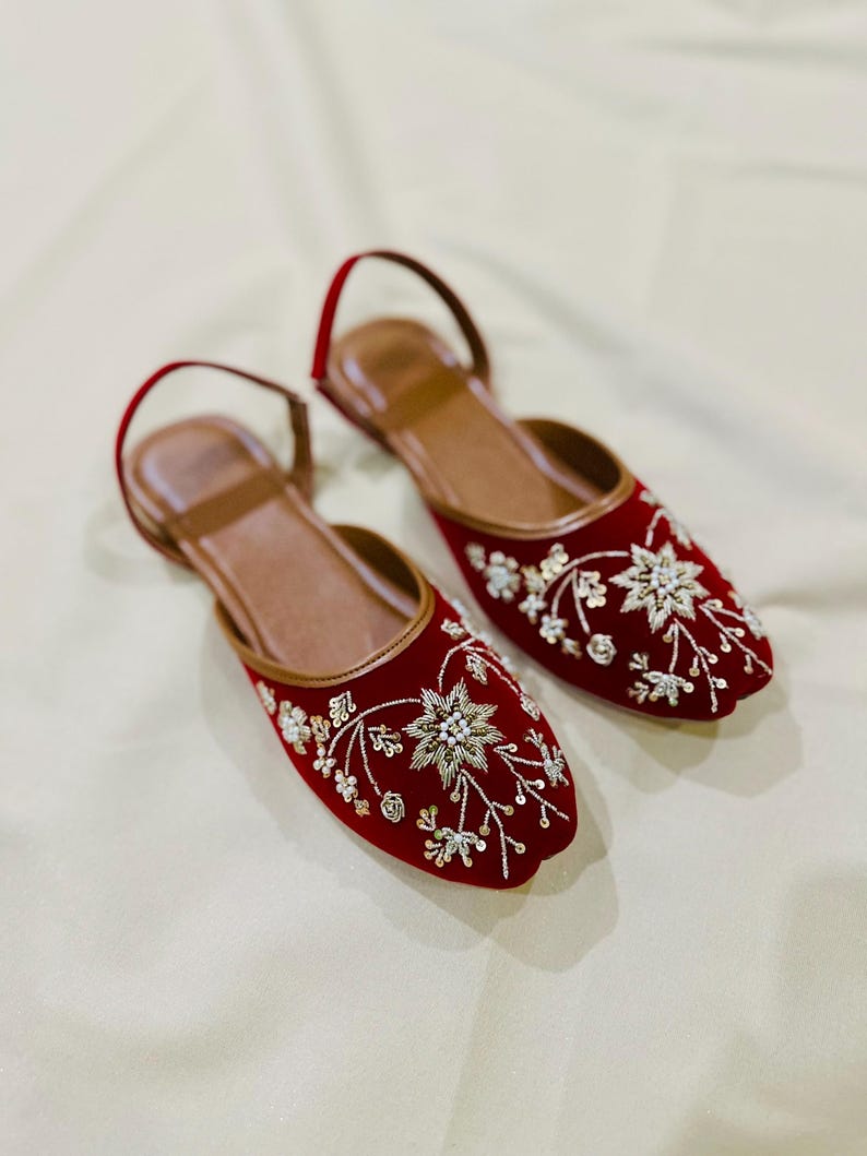 May include: A pair of red velvet flats with intricate floral embroidery in silver and gold. The shoes have a brown leather insole and a thin ankle strap. The flats are a stylish and elegant footwear option.