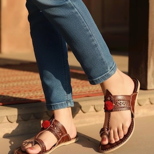 Könnte beinhalten: Braune Ledersandalen mit gewebten und geflochtenen Details, mit einem roten Bommel-Akzent. Die Sandalen haben eine Zehenschlaufe und einen Riemen, der über den Fuß verläuft. Die Sandalen werden mit einer blauen Jeans getragen.