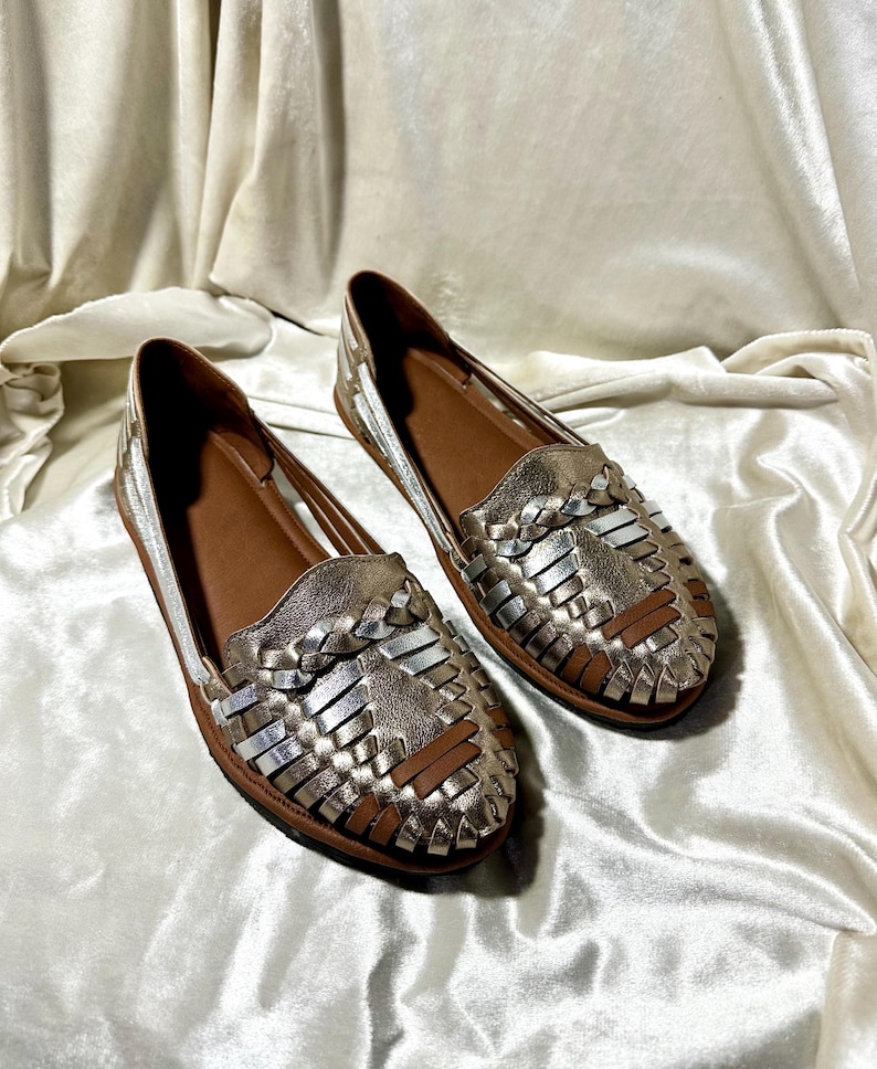 May include: A pair of metallic gold and silver woven leather loafers. The shoes have a brown leather insole and sole. The woven design covers the top of the shoe. The shoes are displayed on a white satin background.