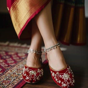 May include: Red velvet flats with intricate gold and silver floral embroidery. The shoes have a rounded toe and a thin, gold-colored trim. Silver anklets with dangling charms are worn with the shoes. The background includes a patterned rug and a red and gold sari.