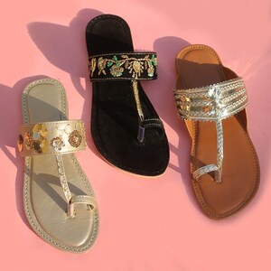 May include: Three pairs of flat sandals in gold, black, and brown, each with unique embellishments. The gold sandal features floral accents, the black sandal has embroidered details, and the brown sandal has silver straps. All have a toe loop design.