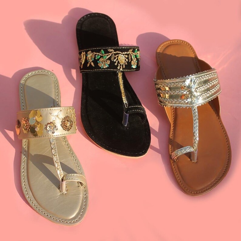 May include: Three pairs of flat sandals in gold, black, and brown, each with unique embellishments. The gold sandal features floral accents, the black sandal has embroidered details, and the brown sandal has silver straps. All have a toe loop design.