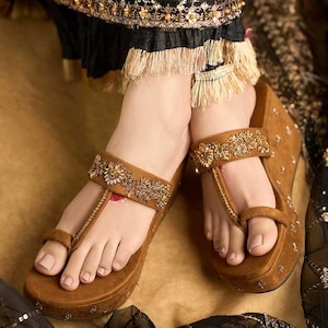 May include: Brown wedge sandals with ornate gold and beige embellishments. The sandals feature a toe loop and ankle strap, with a platform sole. The design includes intricate beadwork and floral patterns, creating an elegant look.