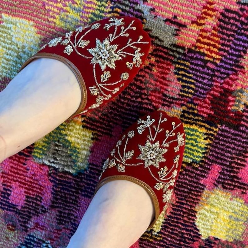 May include: A pair of red velvet slippers with intricate gold and silver floral embroidery. The slippers have a brown leather trim and are displayed on a colorful patterned rug. The design features a central flower with surrounding leaves and smaller floral accents.