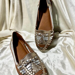 May include: A pair of gold and silver woven leather flats. The shoes have a brown insole and a woven design on the top. The shoes are displayed on a white, textured fabric.