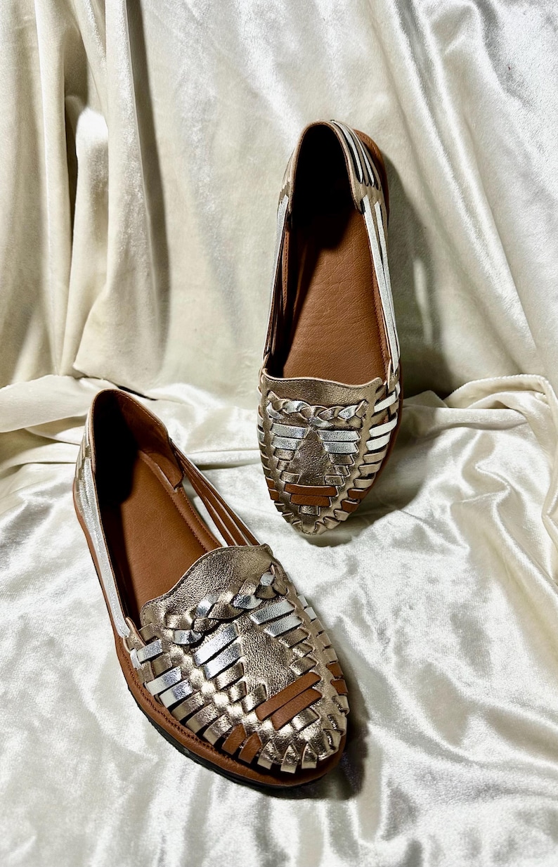 May include: A pair of gold and silver woven leather flats. The shoes have a brown insole and a woven design on the top. The shoes are displayed on a white, textured fabric.