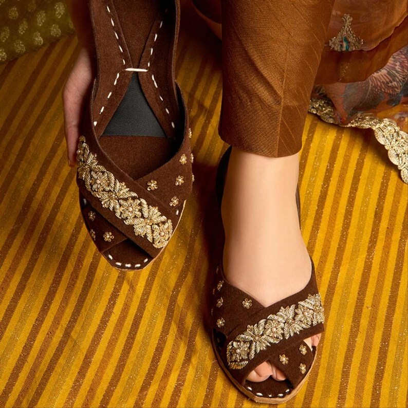 May include: Brown suede slip-on sandals with gold floral embroidery. The sandals have a criss-cross design across the top and are embellished with small gold accents. The soles are a light tan color, and the sandals are on a yellow and brown striped surface.