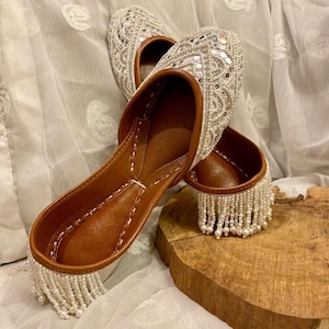 Pode incluir: Um par de sapatos rasos adornados. Os sapatos apresentam interior e sola em couro castanho, com cabedais brancos com contas e espelhados. A guarnição é adornada com uma franja de contas brancas. Os sapatos são exibidos em uma superfície de madeira.