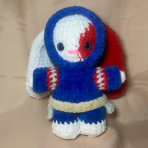 May include: A handmade plush bunny doll with a blue hooded jacket, blue shorts, and white and red accents. The bunny has a white face with red markings, blue eyes, and a pink mouth. The bunny's ears are white and red.