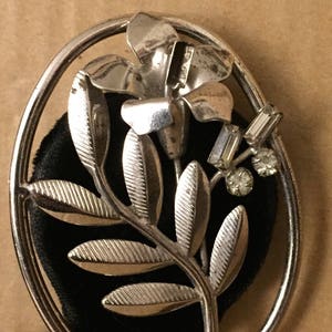 May include: Silver-toned floral brooch with an oval frame. The brooch features a flower and leaf design with clear rhinestone accents. The background is black, and the brooch is set against a neutral-toned surface.