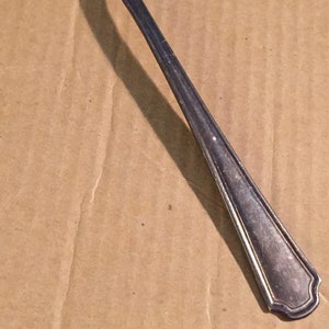 Może przedstawiać: Srebrna łyżka z zaokrągloną miseczką i długim, smukłym trzonkiem. Uchwyt ma dekoracyjny, żebrowany wzór. Łyżka spoczywa na brązowej, teksturowanej powierzchni. Łyżka jest prawdopodobnie do jedzenia.