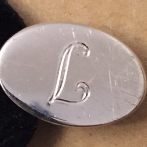 Può includere: Un gemello ovale argentato con la lettera "L" incisa in corsivo. Il gemello ha una finitura lucida. Lo sfondo è di colore neutro. La lettera "L" è in corsivo.