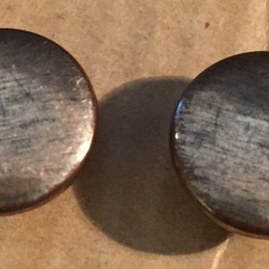 May include: Two circular, metallic buttons with a brushed silver finish and a dark brown edge. The buttons are set against a light brown background, with one casting a shadow.