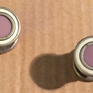 May include: Two vintage cufflinks with a circular design. Each cufflink features a light purple center, surrounded by a white ring and a silver-colored metal frame. The cufflinks are displayed on a brown surface.