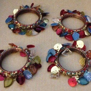 May include: Four decorative bracelets with a brown metal band, adorned with colorful, iridescent shell-shaped accents in shades of red, blue, green, and gold. The bracelets also feature small, colorful beads.