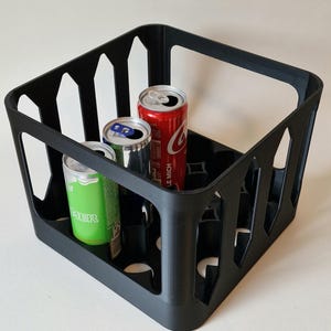 Könnte beinhalten: Ein schwarzer Getränketräger aus Kunststoff mit drei Aluminiumdosen. Eine Dose ist rot mit weißem Text, eine weitere silberfarben und die dritte grün. Der Träger hat eine quadratische Form mit ausgeschnittenen Designs an den Seiten.
