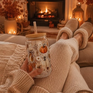 May include: A clear glass tumbler with a bamboo lid and a clear straw, featuring a printed design of orange and white pumpkins, small white daisies, brown checkered bows, and tiny gold stars scattered across the surface of the cylindrical glass.
