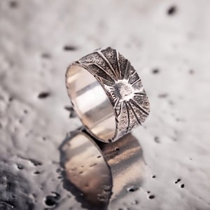 Könnte beinhalten: Ein silberner Ring mit einem strukturierten Design, das an einen Sonnenstrahl erinnert. Der Ring ist breit und hat ein poliertes Innenleben.