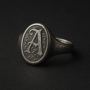 Könnte beinhalten: Ein silberner Siegelring mit einer ovalen Fläche, die einen gravierten Buchstaben 'A' zeigt, umgeben von einem dekorativen floralen Design.