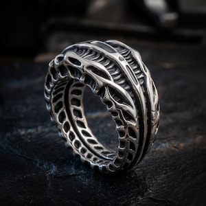 May include: A detailed, silver-colored ring with an intricate, textured design. The ring features a series of interconnected, organic-looking shapes, creating a unique and eye-catching pattern. The ring's design is reminiscent of a biomechanical or alien aesthetic.