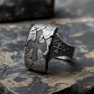 Könnte beinhalten: Silberfarbener Ring mit einer großen, strukturierten Oberfläche mit einem Kreuzdesign. Der Ring hat ein rissiges, gealtertes Aussehen mit aufwendigen Details am Band und am Kreuz. Der Ring liegt auf einer dunklen, strukturierten Oberfläche.