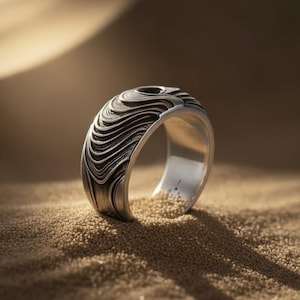 Könnte beinhalten: Ein silberner Ring mit einem einzigartigen wellenartigen Muster. Der Ring ist vor einem sandigen Hintergrund platziert, der sein kompliziertes Design hervorhebt. Die Oberfläche des Rings hat ein strukturiertes Aussehen, was seine visuelle Attraktivität erhöht. Der Ring ist ein Schmuckstück.