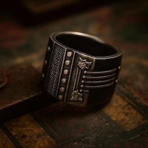 Op de afbeelding: Een brede, donkere metalen ring met ingewikkelde details. De ring heeft een getextureerd chevronpatroon, kleine cirkelvormige accenten en gestileerde wolfskopontwerpen. Het algehele ontwerp heeft een vintage of gotische esthetiek.