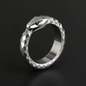 Puede incluir: Un anillo de plata con forma de serpiente con un ojo de diamante negro. El anillo está hecho de metal facetado y tiene un diseño geométrico.