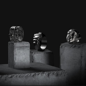 Peut inclure: Trois bagues argentées exposées sur des blocs de béton gris sur fond noir. Les bagues présentent des designs uniques : l'une avec un aspect boucle, une autre avec un motif géométrique et une troisième avec un visage sculpté.
