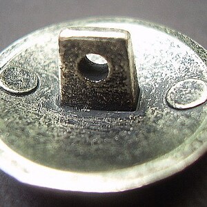 May include: Close-up of a silver-toned button with a square loop and a circular hole. The button has a textured surface and two small, oval-shaped details on the rim. The button is likely for clothing or other fabric items.