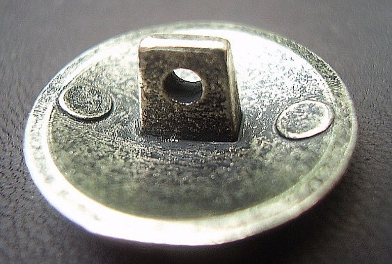 May include: Close-up of a silver-toned button with a square loop and a circular hole. The button has a textured surface and two small, oval-shaped details on the rim. The button is likely for clothing or other fabric items.