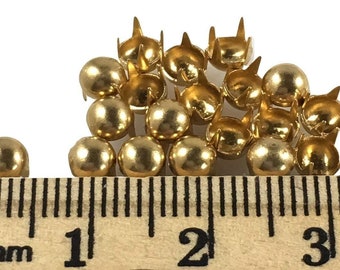 May include: A close-up of a pile of gold-colored metal studs with a ruler showing 1 inch in length. The studs have a round head and four prongs.