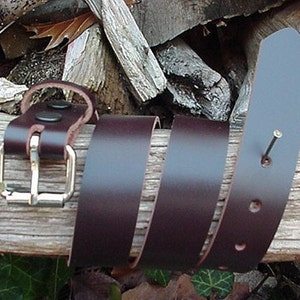 May include: A brown leather belt with a silver buckle and multiple holes. The belt is coiled on a wooden surface.