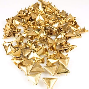 May include: A large pile of gold-colored triangular metal studs. The studs have a pointed tip and a flat base. They are commonly used for decorating clothing, bags, and other accessories.