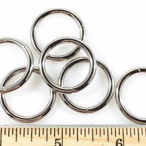 Puede incluir: Seis anillos de metal plateado, de aproximadamente 2,5 cm de di&aacute;metro, est&aacute;n esparcidos sobre una superficie blanca. Una regla de madera con marcas en pulgadas es visible en la parte inferior de la imagen.