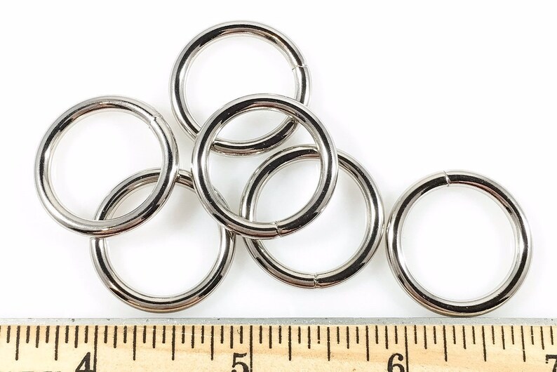 Puede incluir: Seis anillos de metal plateado, de aproximadamente 2,5 cm de di&aacute;metro, est&aacute;n esparcidos sobre una superficie blanca. Una regla de madera con marcas en pulgadas es visible en la parte inferior de la imagen.