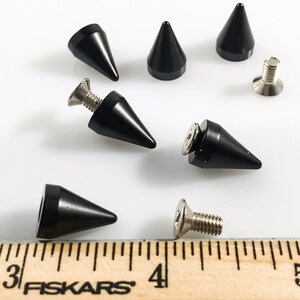 May include: A set of eight black metal pyramid studs with silver screws. The studs are approximately 2.5 cm in length. The studs are shown on a wooden ruler with the text "Fiskars" printed on it.