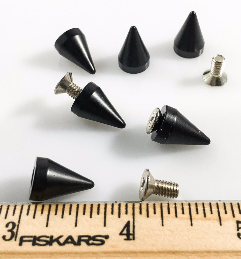 May include: A set of eight black metal pyramid studs with silver screws. The studs are approximately 2.5 cm in length. The studs are shown on a wooden ruler with the text "Fiskars" printed on it.